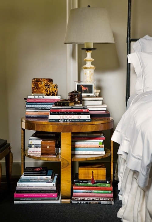bedside table with top and shelves filled with stacks of books, trinkets, and a lamp
