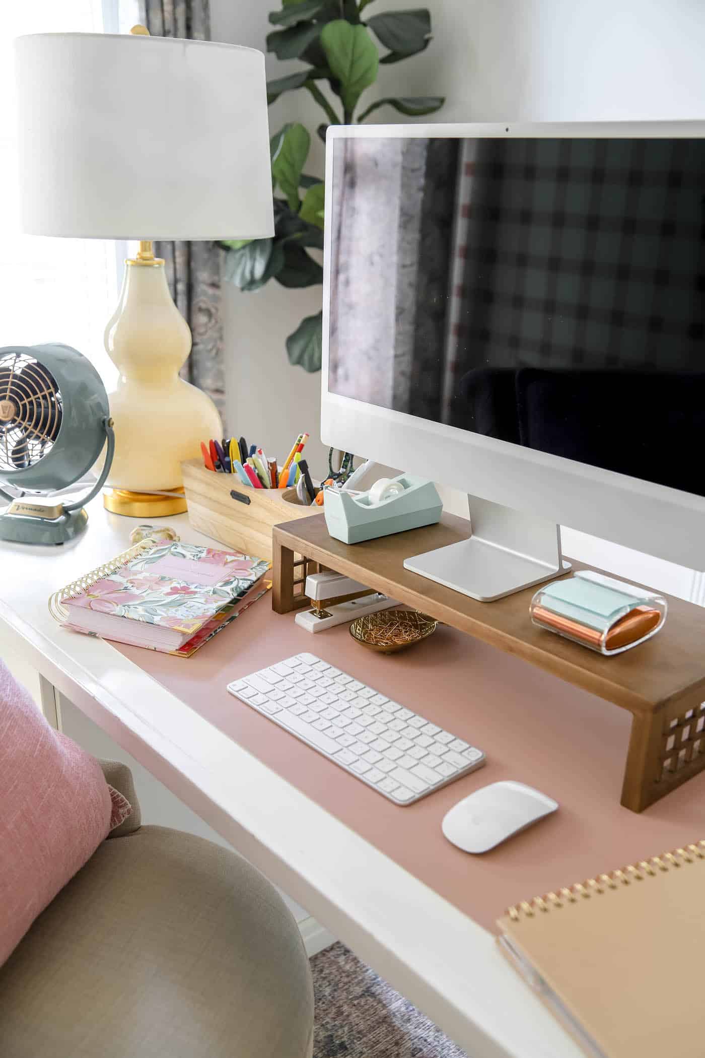 organized desk space with mac computer and desk riser, fan and lamp.