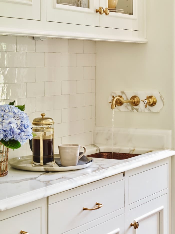 white butler's pantry with subway tile backsplash, marble countertop, brasss faucet and small copper sink.