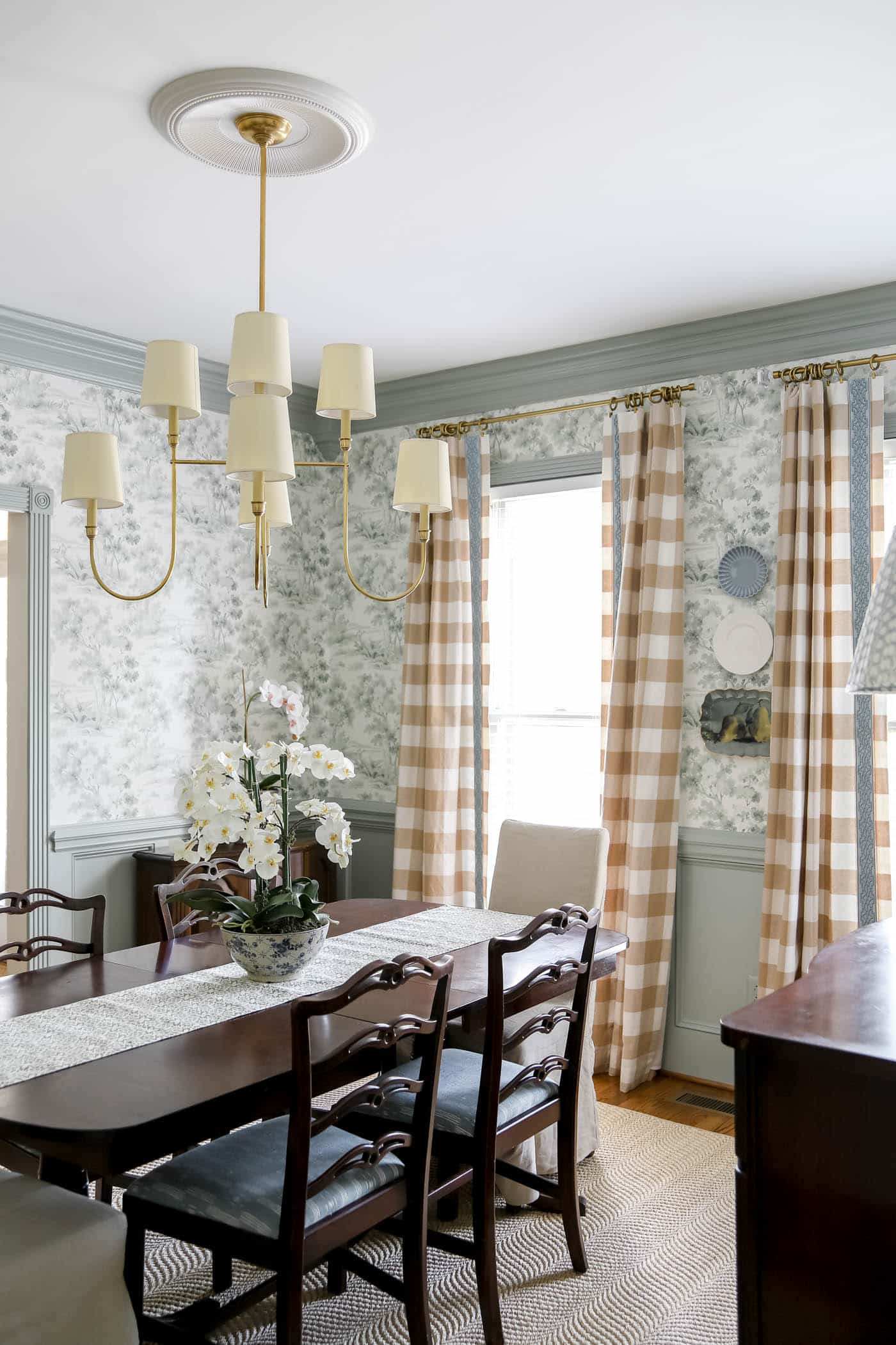 dining room with plates hung on a wall, gingham curtains, brass chandelier and dark brown dining table and chairs.