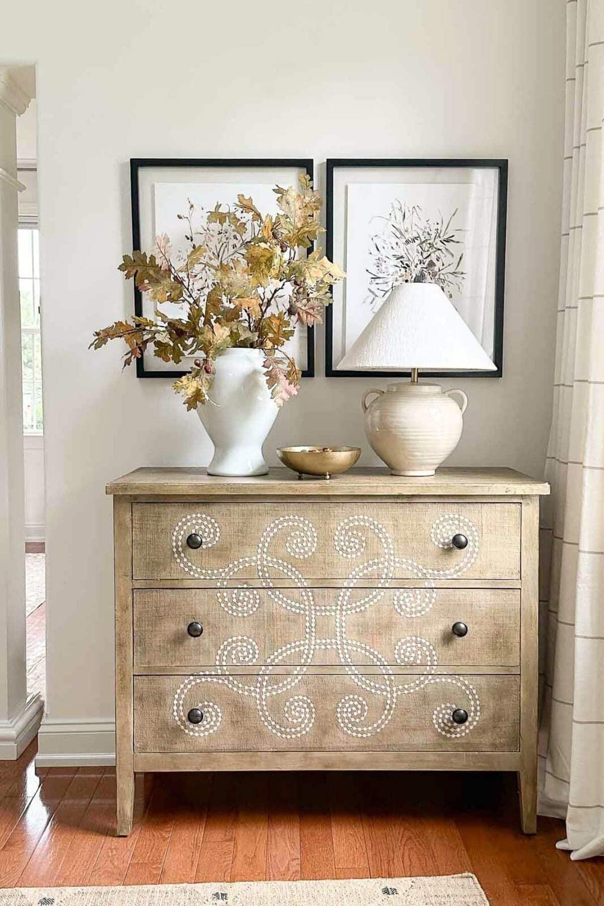 light wood dresser with fall foliage in a vase and a lamp.