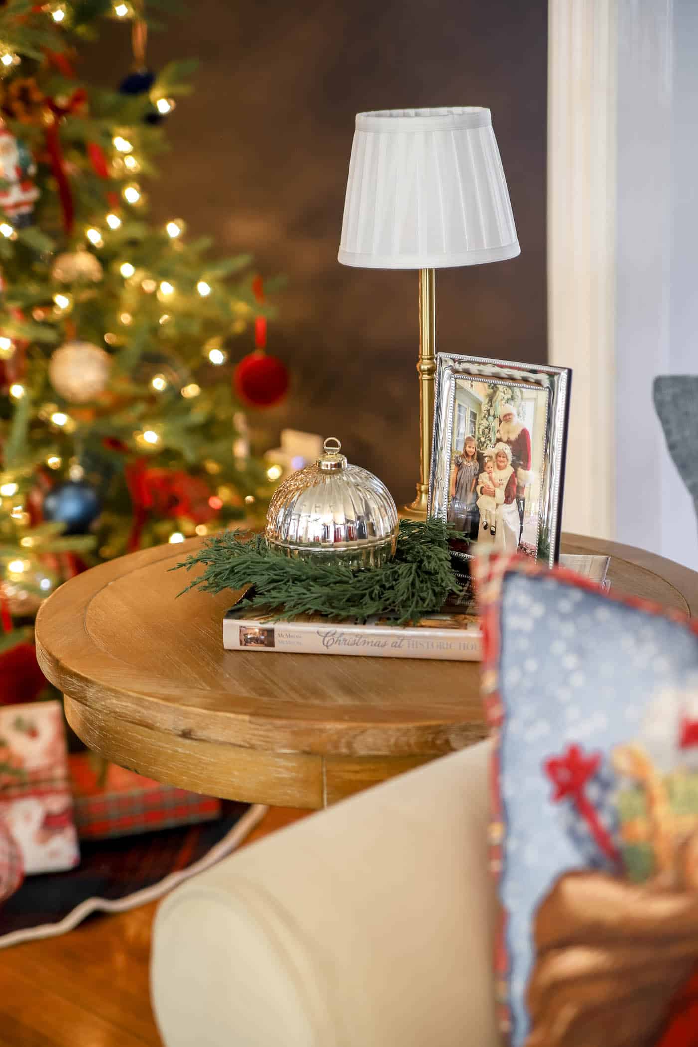 side table with Santa and children in a frame, small cordless lamp and ornament candle with a christmas coffee table book and greenery.