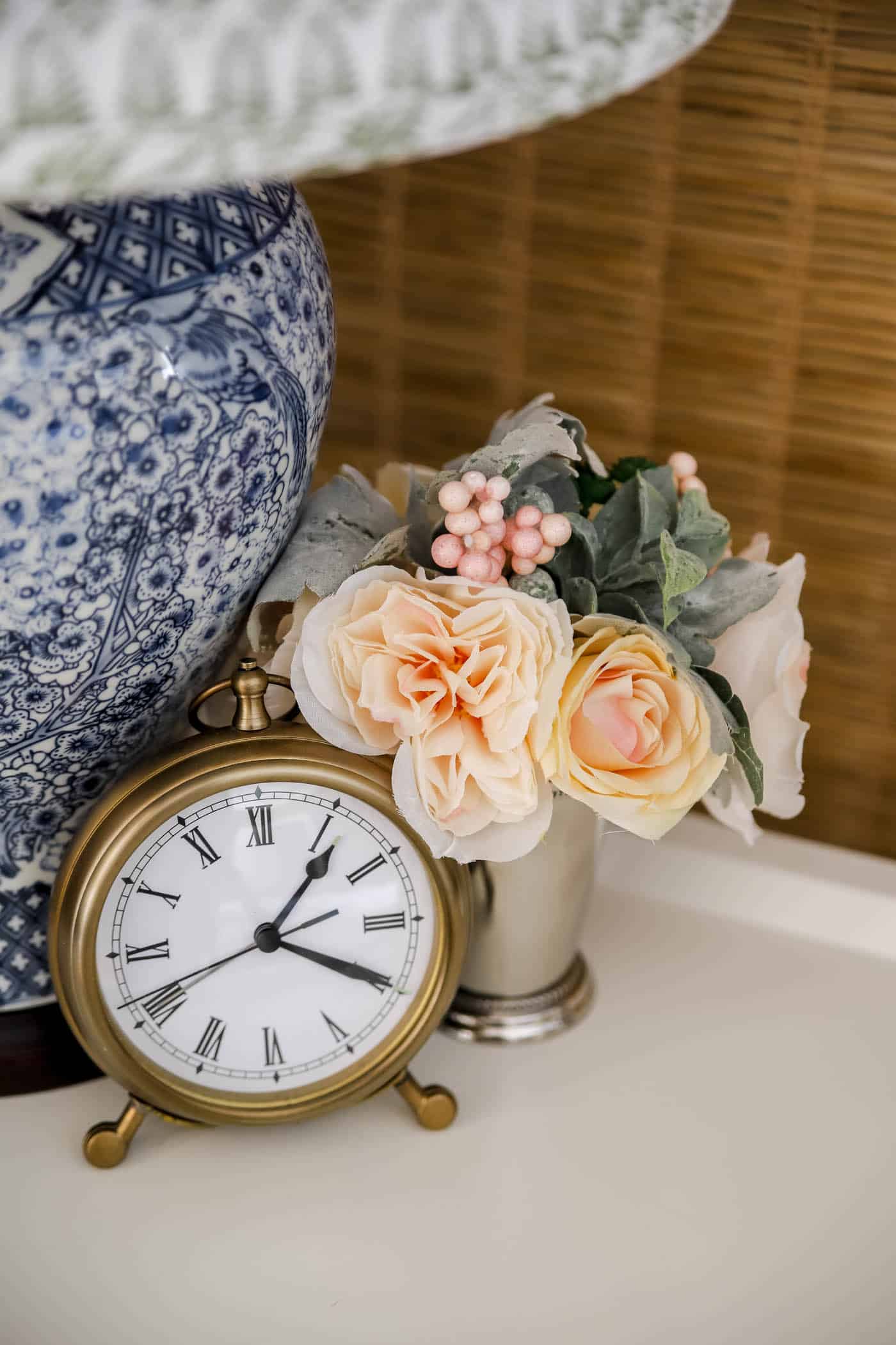small bouquet of faux flowers in a mint julep cup, along with a pocket watch clock and blue and white lamp vase on a nightstand. 