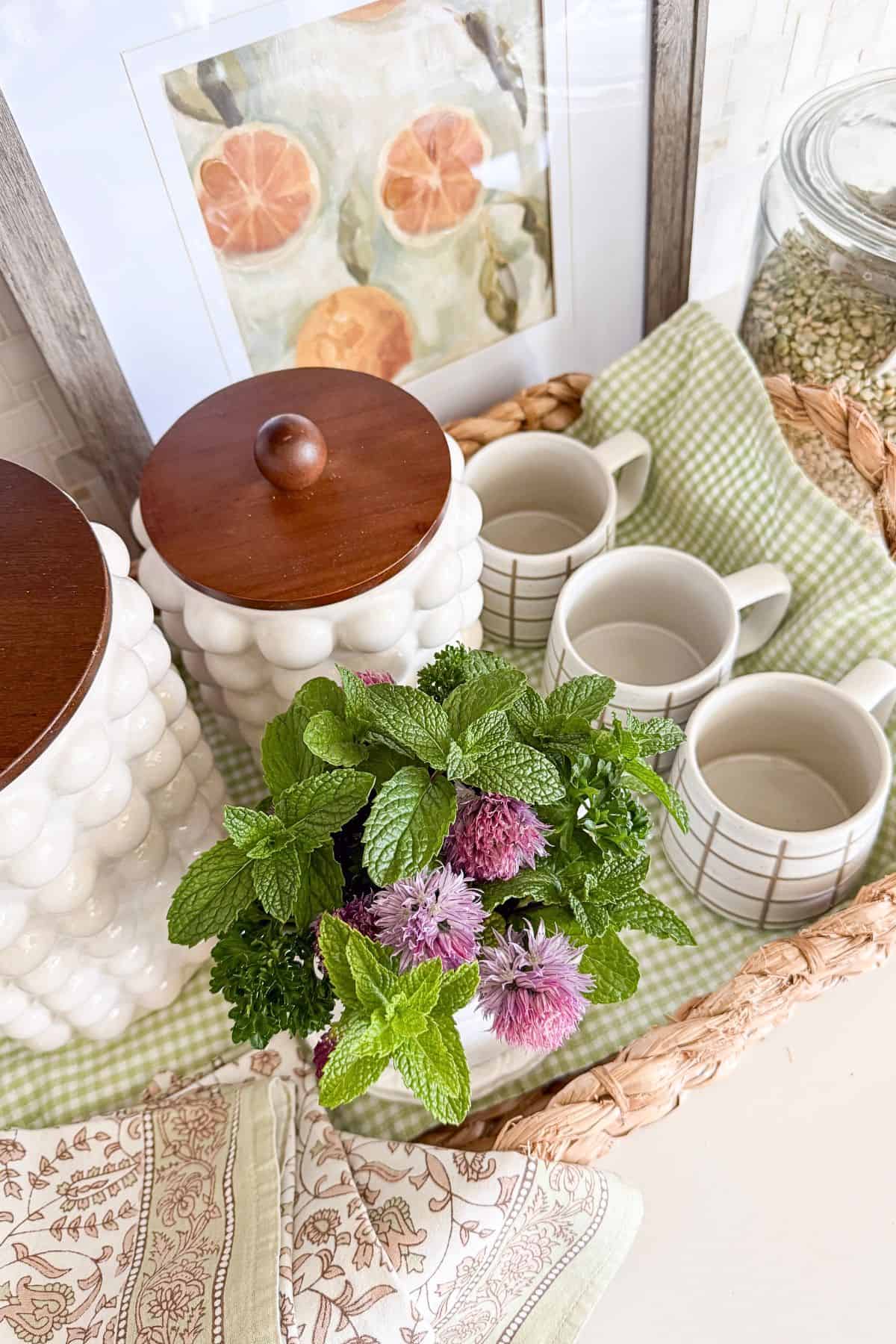 Summer coffee bar vignette with green check runner, chive herbs, blossoms, mugs and baskets. 